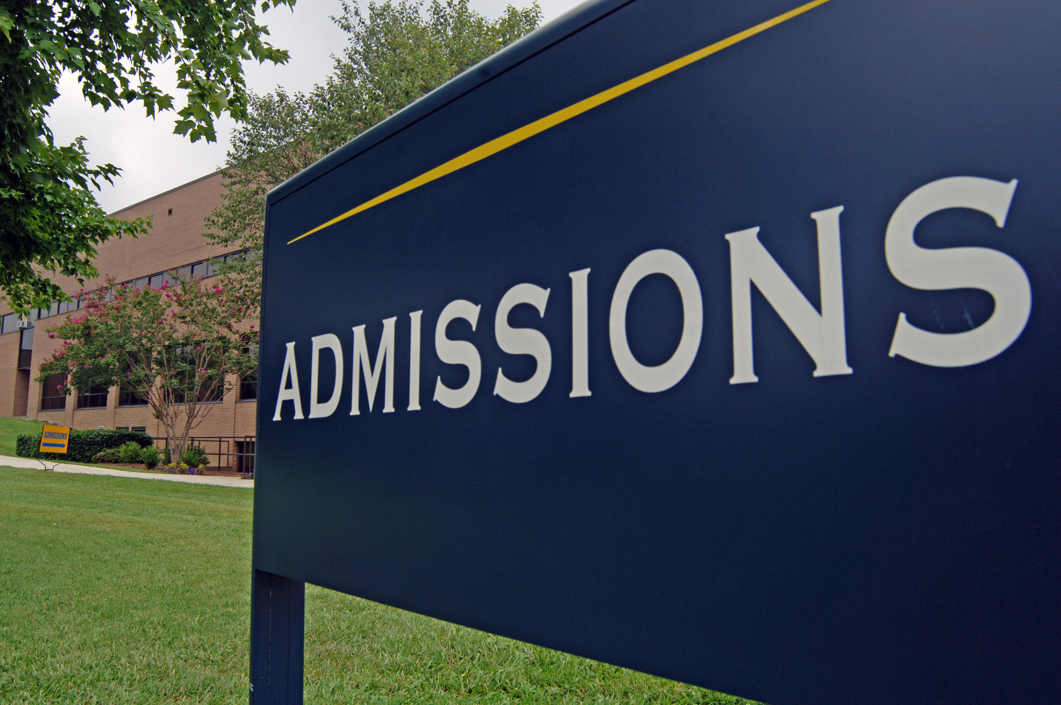A big blue sign with white letters saying "admissions" in the foreground with grass and college like building in the background.